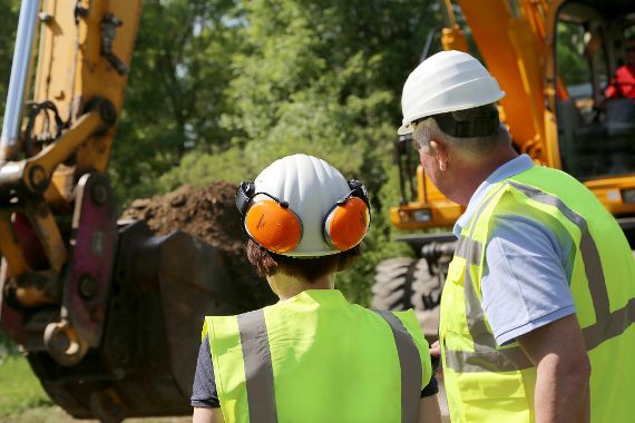 Foto von zwei Personen, die mit Bauhelm auf einer Baustelle einen Bagger beobachten